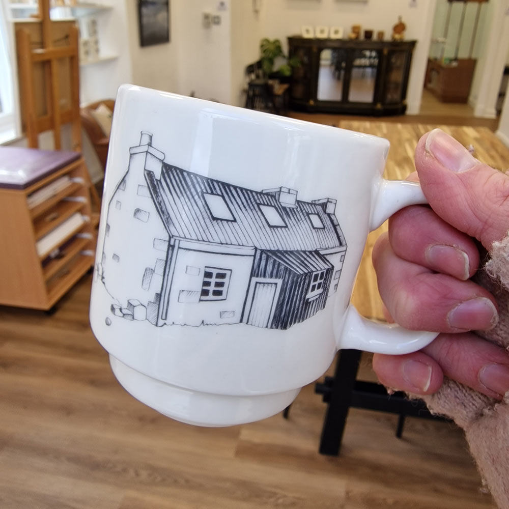Hand holding a small white mug with the picture of a stone cottage on it